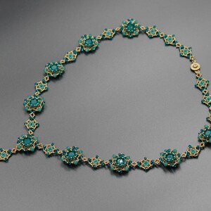 Pu&ograve; includere: Una collana artigianale con perline color teal e oro. La collana presenta un design floreale con pi&ugrave; gruppi di perline teal collegati da maglie color oro. La chiusura &egrave; dorata. La collana &egrave; esposta su uno sfondo grigio.