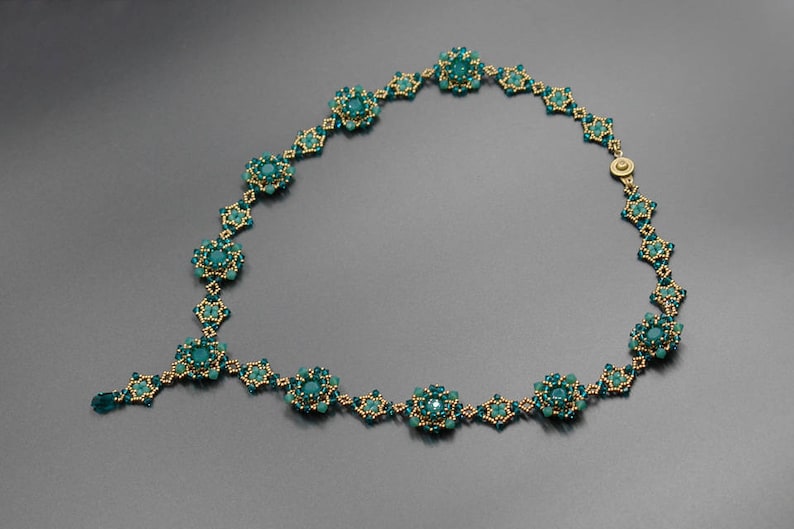 Pu&ograve; includere: Una collana artigianale con perline color teal e oro. La collana presenta un design floreale con pi&ugrave; gruppi di perline teal collegati da maglie color oro. La chiusura &egrave; dorata. La collana &egrave; esposta su uno sfondo grigio.