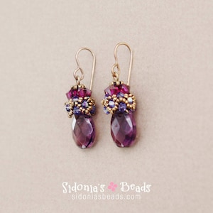Può includere: Un paio di orecchini viola e dorati con un cristallo a forma di lacrima e un design a perline.