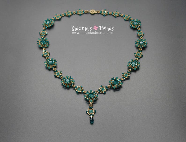 Pu&ograve; includere: Collana artigianale con perline color verde acqua e oro. La collana presenta un design floreale con un pendente centrale e una catena delicata. Il testo "Sidonia's Beads" e l'indirizzo del sito web sono visibili.