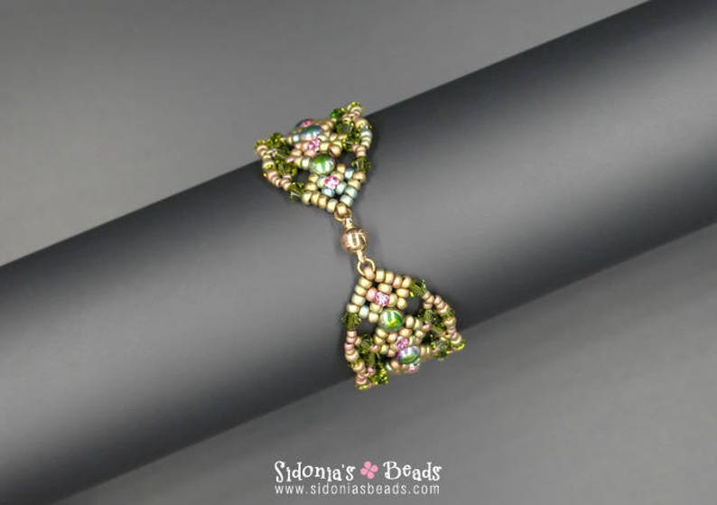 May include: A green, pink and gold beaded bracelet with a geometric design. The bracelet is made with seed beads and features a delicate, intricate pattern.