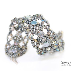 May include: A silver and grey beaded bracelet with a woven design. The bracelet features a variety of beads, including small round beads, larger hexagonal beads, and clear crystal beads.