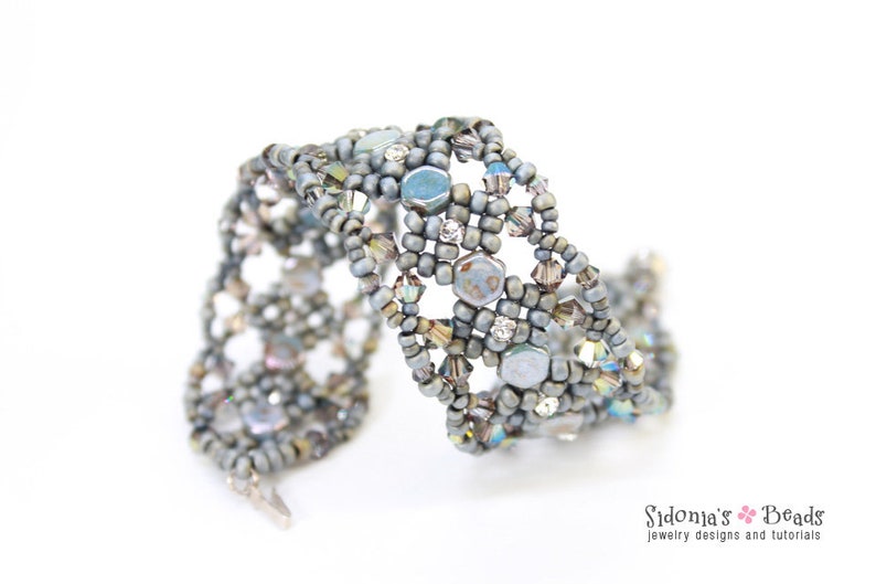 May include: A silver and grey beaded bracelet with a woven design. The bracelet features a variety of beads, including small round beads, larger hexagonal beads, and clear crystal beads.