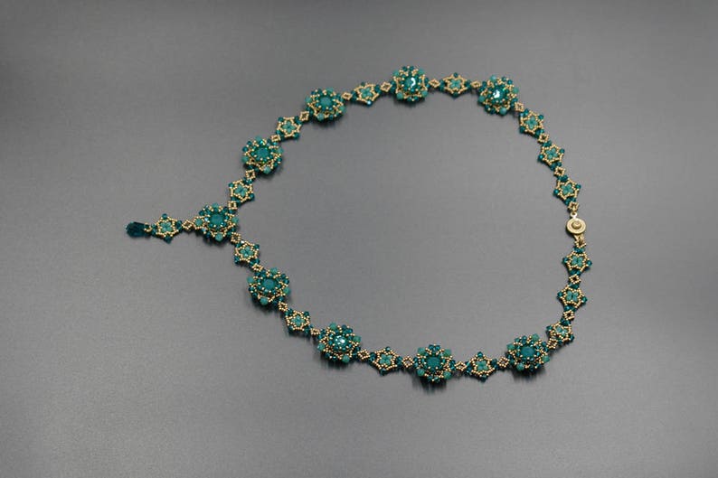 Pu&ograve; includere: Una collana artigianale con perline color teal e dettagli in metallo dorato. La collana presenta un design floreale con un pendente centrale e una catena delicata. La collana &egrave; esposta su una superficie grigio scuro.