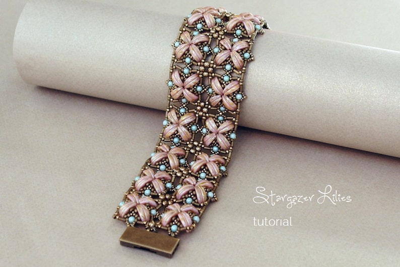 Pu&ograve; includere: Un braccialetto di perline con un design floreale rosa e blu. Il braccialetto &egrave; realizzato con piccole perline a seme e ha una chiusura in metallo. Il braccialetto &egrave; su una superficie marrone chiaro. Il testo "Stargazer Lilies tutorial" &egrave; visibile nell'immagine.