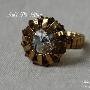 Könnte beinhalten: Ein goldfarbener Ring mit einem großen, facettierten, klaren Mittelstein. Der Ring ist mit kleinen, rechteckigen Goldperlen und braunen Akzenten verziert, mit dem Text "Half Tila Ring". Der Ring hat ein dehnbares Band.