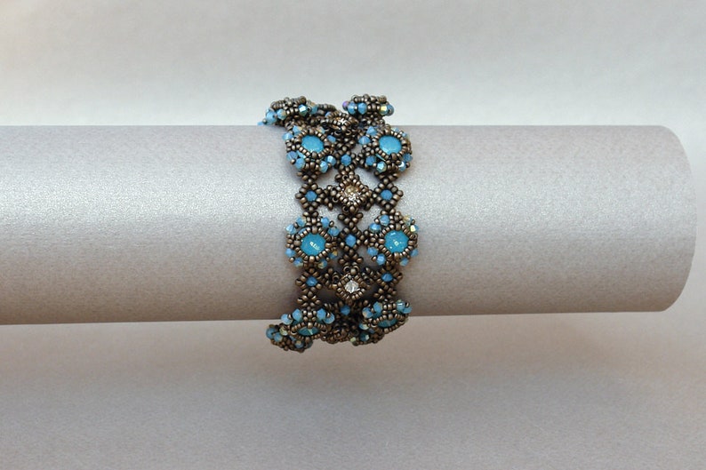 K&ouml;nnte beinhalten: Ein Armband mit Perlen in geometrischem Design mit blauen und klaren Perlen. Das Armband ist aus bronzefarbenen Perlen gefertigt und auf einer wei&szlig;en Oberfl&auml;che abgebildet.