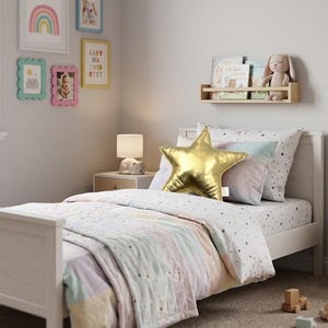May include: A white wooden bed frame featuring a quilt with pastel stripes and a star-patterned sheet. A metallic gold star-shaped pillow rests on the bed alongside a plush rabbit toy on a wall-mounted shelf. A small lamp sits on a bedside table.