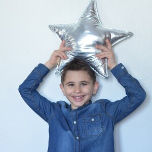 May include: A shiny, metallic silver star-shaped pillow with a smooth, reflective surface. The star has five pointed tips and a plush, stuffed construction, creating a soft and rounded appearance across its entire form.