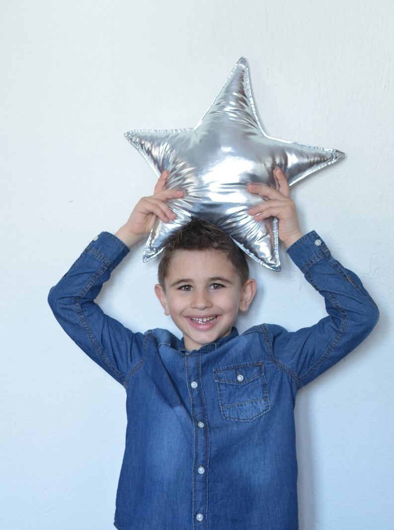 May include: A shiny, metallic silver star-shaped pillow with a smooth, reflective surface. The star has five pointed tips and a plush, stuffed construction, creating a soft and rounded appearance across its entire form.