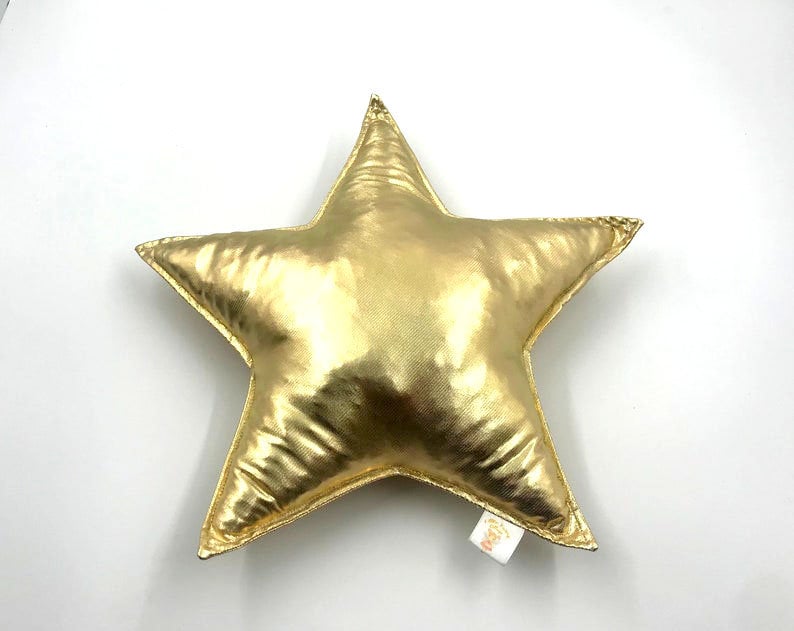 May include: A star-shaped pillow with a metallic gold finish. The pillow features a smooth, reflective surface with visible stitched seams along the edges and a small white fabric tag attached to the lower right point of the star shape.