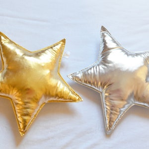 May include: Two star-shaped pillows with a metallic finish. One pillow is gold and the other is silver. Both pillows feature a smooth, reflective surface and stitched edges, creating a distinct five-pointed star silhouette with a soft, plush appearance.