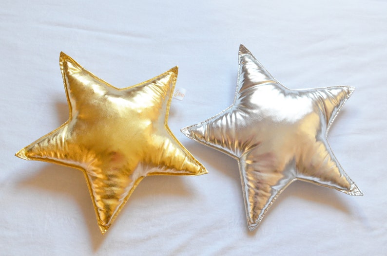 May include: Two star-shaped pillows with a metallic finish. One pillow is gold and the other is silver. Both pillows feature a smooth, reflective surface and stitched edges, creating a distinct five-pointed star silhouette with a soft, plush appearance.