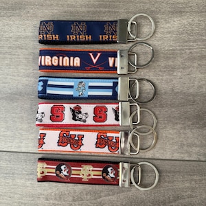 May include: Six fabric keychains with metal rings. The top is navy with gold Notre Dame text. Second is navy with orange Virginia text. Third is blue and white stripes with a mascot. Fourth is white with red NC State logos. Fifth is white with orange Syracuse text. Bottom is red and gold with Florida State logos.