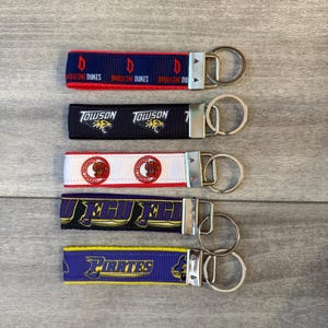 May include: Five fabric keychains with metal rings. The first is navy with red Duquesne Dukes text. The second is black with Towson text and a tiger logo. The third is white with red circular logos. The fourth is purple with ECU text. The fifth is purple with yellow Pirates text.