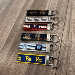 May include: A collection of fabric keychains with metal key rings. The keychains feature various college team logos and text, including "Wake Forest", "Georgia Tech", "Virginia", "Notre Dame", "North Carolina", and "Pitt". The keychains are in various colors.