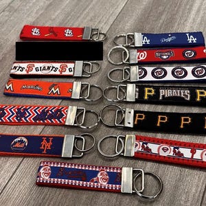May include: Collection of fabric keychains featuring various Major League Baseball team logos and colors. Keychains include teams like the Dodgers, Nationals, and Pirates, with silver-tone hardware.