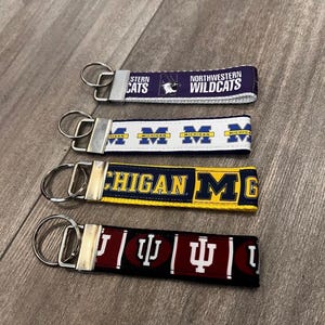 May include: Four fabric keychains with silver metal key rings. The keychains feature the text "NORTHWESTERN WILDCATS", "MICHIGAN", and "MICHIGAN" in various colors. The fourth keychain has the text "I" and a symbol.