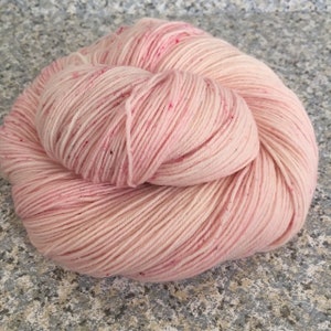 May include: A skein of soft pink yarn with subtle variations in color and tiny flecks of darker pink. The yarn is tightly wound, showing the individual strands. Ideal for knitting or crocheting projects.