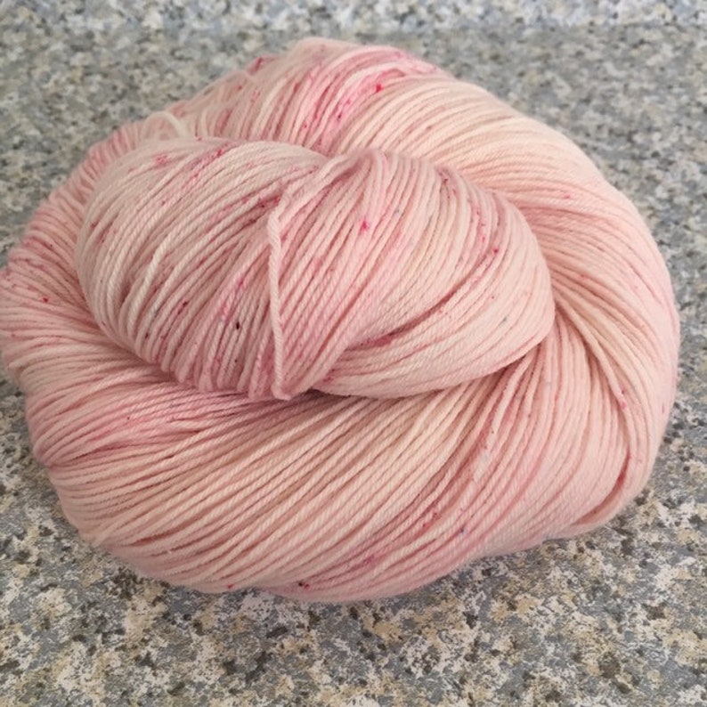 May include: A skein of soft pink yarn with subtle variations in color and tiny flecks of darker pink. The yarn is tightly wound, showing the individual strands. Ideal for knitting or crocheting projects.