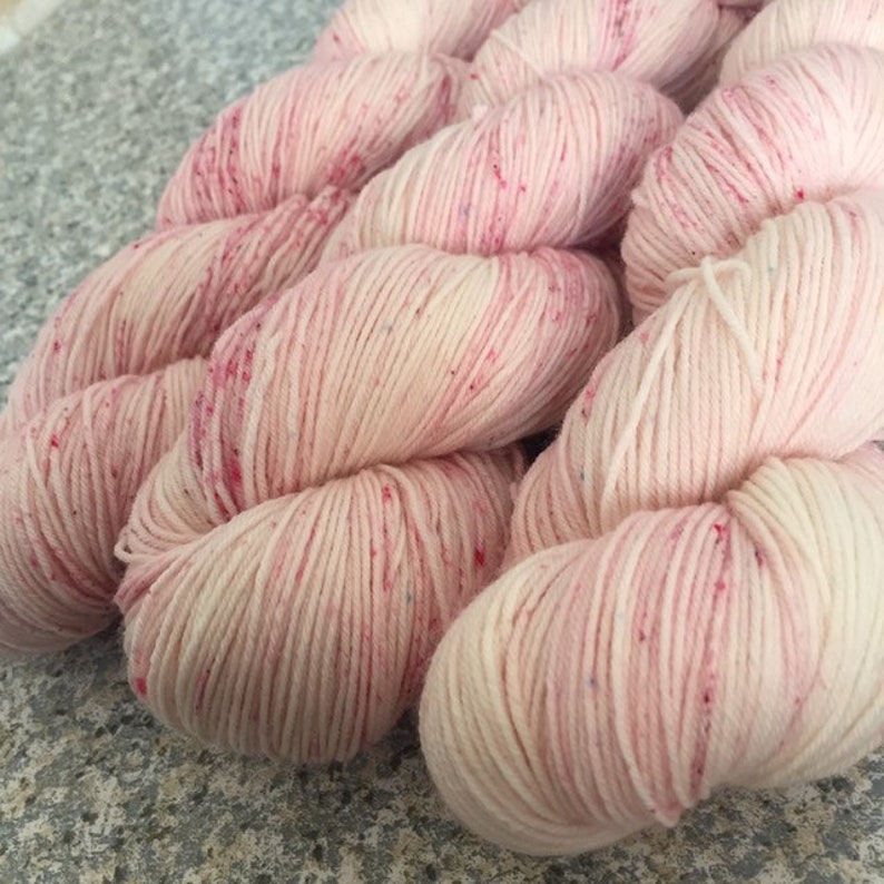 May include: Close-up of several skeins of pink yarn with red speckles. The yarn is tightly wound and appears soft, ideal for knitting or crocheting projects. The color is a light, pastel pink.