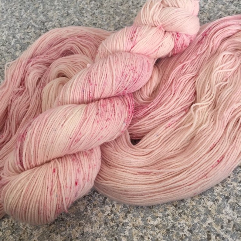 May include: Close-up of pink yarn skeins. The yarn has a speckled appearance with darker pink and red flecks throughout. The yarn is soft and appears to be made of natural fibers, suitable for knitting or crocheting projects. The color is a soft, muted pink.