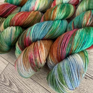 Puede incluir: Primer plano de varios ovillos de hilo teñido a mano en varios colores. El hilo presenta una mezcla de verdes, azules, rojos y marrones. El hilo está dispuesto sobre una superficie de madera de color claro.