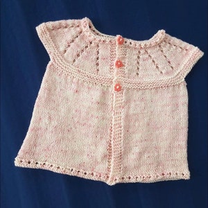 May include: A light pink knitted baby top with a round collar and three flower-shaped buttons. The top has a delicate, openwork design on the collar and sleeves. The yarn has flecks of pink throughout. The garment is displayed on a dark blue background.