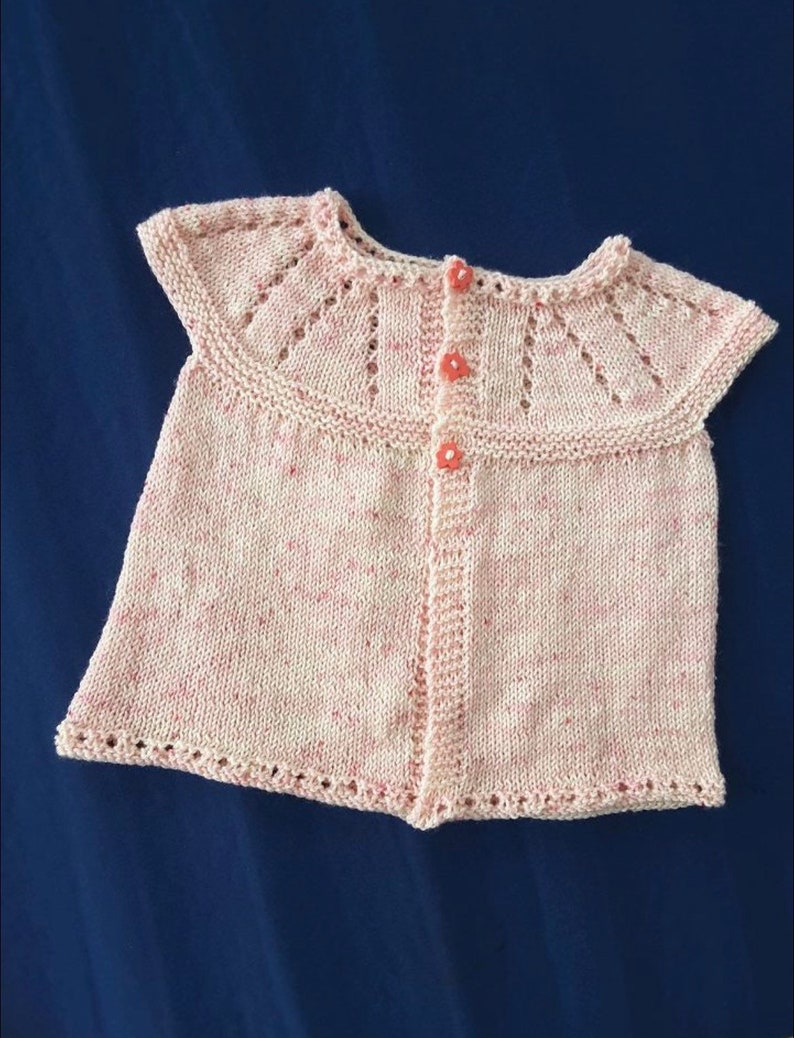 May include: A light pink knitted baby top with a round collar and three flower-shaped buttons. The top has a delicate, openwork design on the collar and sleeves. The yarn has flecks of pink throughout. The garment is displayed on a dark blue background.