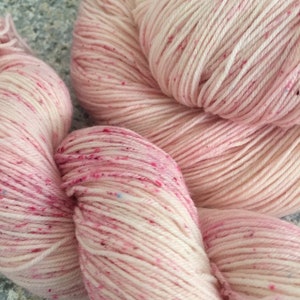May include: Close-up of several skeins of yarn in shades of pink and cream. The yarn has a speckled appearance with flecks of red and blue. The yarn is tightly wound and appears soft, ideal for knitting or crochet projects.