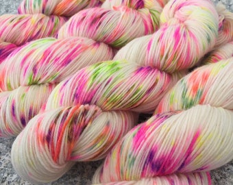 Puede incluir: Un ovillo de hilo blanco con variaciones de color rosa, naranja, verde y morado. El hilo está teñido a mano y tiene un aspecto moteado.