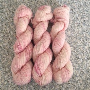 May include: Three skeins of hand-dyed yarn in a soft pink hue with darker pink speckles. The yarn is twisted into a rope-like shape, showcasing its texture and color variations. Ideal for knitting and crochet projects.