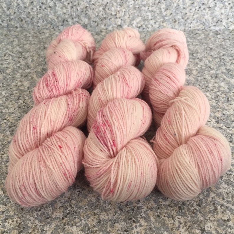 May include: Several skeins of light pink yarn with flecks of red and pink. The yarn is arranged in a cluster, showing the texture and color variations. Ideal for knitting and crochet projects.