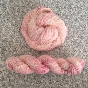May include: Two skeins of pink yarn. One is a large, wound ball, and the other is a twisted hank. The yarn has a speckled appearance with darker pink flecks throughout the light pink and cream-colored fibers. Ideal for knitting or crochet projects.