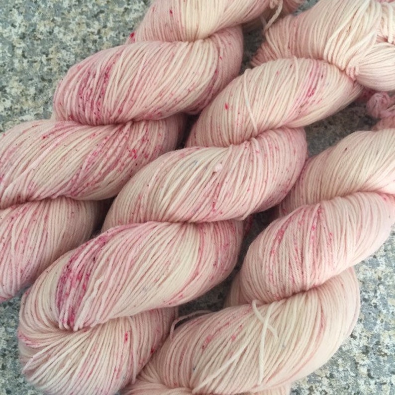 May include: Close-up of several skeins of yarn. The yarn is a light pink color with darker pink speckles throughout. The yarn is tightly wound and appears soft, ideal for knitting or crocheting projects. The background is a neutral gray.