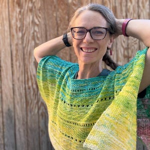 May include: A hand-knitted shawl in shades of yellow, green, and blue. The shawl has a textured, open-weave design with a gradient effect. The woman in the image is wearing glasses and has her hands behind her head.