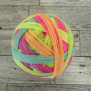 Puede incluir: Ovillo de lana multicolor con hilos amarillo neón, rosa, naranja y turquesa. La lana está enrollada apretadamente, creando una apariencia texturizada. Ideal para proyectos de punto o ganchillo. Diámetro aproximado de 10 cm.
