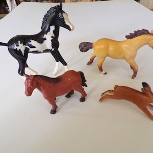 May include: A collection of four animal figurines including a black and white pinto horse, a tan horse with a dark mane, a solid brown horse, and a brown rabbit in a leaping pose. These small plastic toys are arranged together on a flat surface.