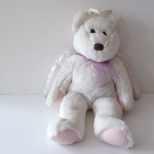Peut inclure: Ours en peluche blanc portant un halo et des ailes violettes. L'ours a un nez brun et des coussinets roses.