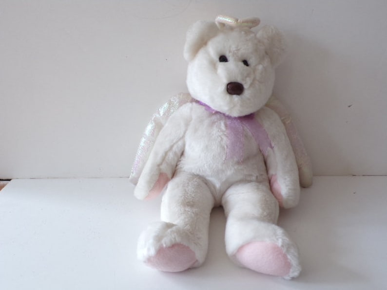 Peut inclure: Ours en peluche blanc portant un halo et des ailes violettes. L'ours a un nez brun et des coussinets roses.