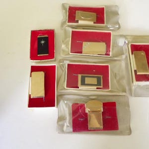 May include: A collection of seven gold-tone metal money clips, each presented in a white box with a red velvet insert. Some clips feature black enamel accents or star motifs, and several are individually sealed in clear plastic protective packaging.