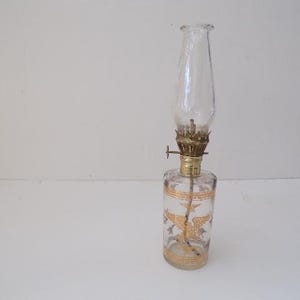May include: A clear glass oil lamp with a gold-coloured metal top and a gold-coloured design on the bottom of the glass. The lamp is on a white surface.