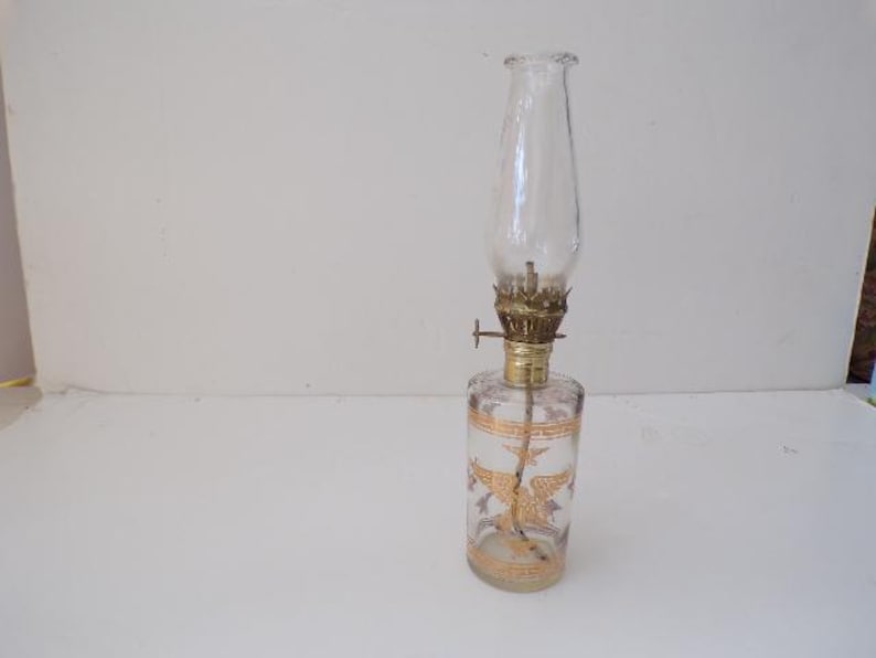 May include: A clear glass oil lamp with a gold-coloured metal top and a gold-coloured design on the bottom of the glass. The lamp is on a white surface.