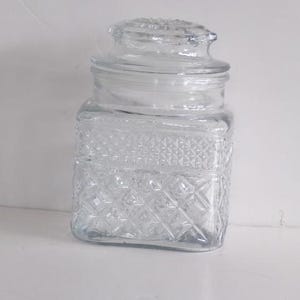 May include: A clear, square glass jar with a lid. The jar features a textured, diamond-patterned design on its body. The lid is also clear and has a decorative knob. This jar is suitable for storing small items.