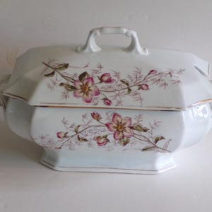 May include: White porcelain serving dish with lid, featuring a floral design in pink and green. The dish has a gold trim and a handle on the lid. The overall shape is octagonal.