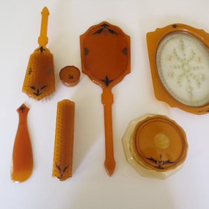 May include: A vanity set in amber-coloured material featuring a hand mirror, a hairbrush, a comb, a small circular container, a larger lidded jar, and an oval tray with a white floral inlay. Each piece is accented with dark, pointed leaf-like motifs.