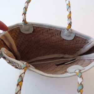 May include: A white woven straw tote bag with a braided leather handle. The handle is white, yellow, and blue.