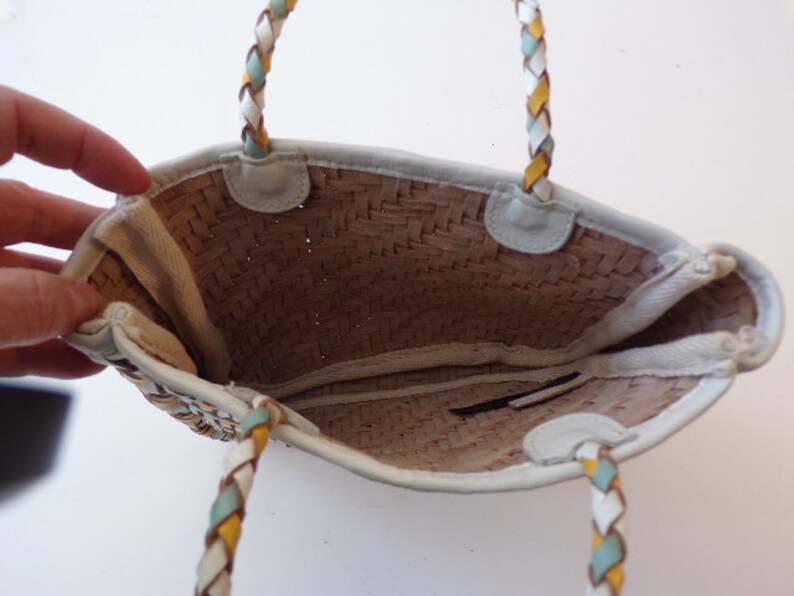 May include: A white woven straw tote bag with a braided leather handle. The handle is white, yellow, and blue.