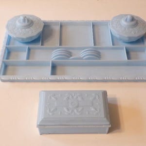 May include: A light blue vintage vanity set with a tray, two lidded dishes, and a rectangular box. The tray has multiple compartments and the lidded dishes and box have floral designs.