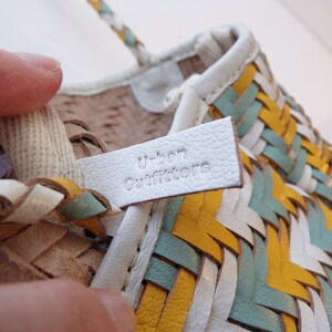 May include: White leather sandals with a woven design in yellow, blue, and white. The sandals have a white leather label that says "Urban Outfitters".